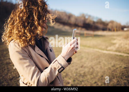 Junge Frau mit dem lockigen Haar zu tragen Rot beige Mantel mit Ihrem Smart Phone stehen draußen in Gelb sonnenbeschienenen Feld auf warmen Herbsttag. Stockfoto