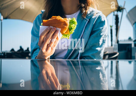 Weibliche Mittagessen im sonnigen Tag. Close-up junge Frau mit Gebissen Hamburger am Tisch sitzen im Café im Freien. Stockfoto