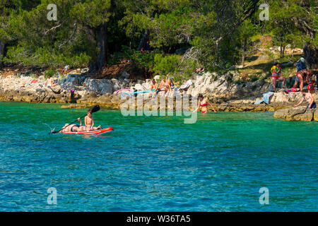 Vater und Sohn paddeln entlang der kroatischen Küste in der Nähe von Malinska auf der Insel Krk, Kroatien, mit reichlich Sonnenanbeter im Hintergrund Stockfoto