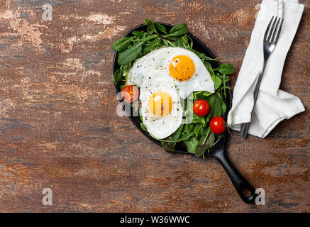 Pfanne mit Spiegelei und Cherry Tomaten und grüne Salate am braunen Hintergrund. Serviette und die Gabel in der Nähe. Konzept der gesunden Ernährung. Flach, Ansicht von oben, kopieren Stockfoto