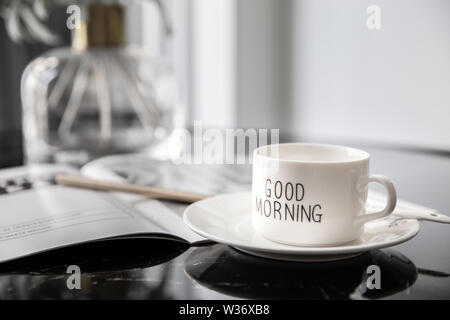 Weiß Kaffeebecher mit Blumenstrauß Maiglöckchen und Notizen 'Guten Morgen' auf Schwarz moderner Marmortisch. Strahlender Sonnenschein morgen refreshm Stockfoto