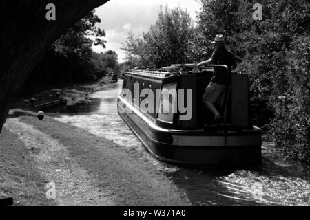 Ein schmales Boot, das von einem Mann mit Sonnenhut auf dem Grand Union Canal direkt nach dem Verlassen einer abgesenkten Schleuse in der Nähe von Aylesbury, Buckinghamshire, gelenkt wird. Stockfoto