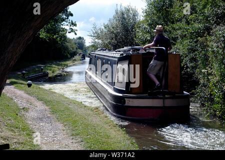 Ein schmales Boot, das von einem Mann mit Sonnenhut auf dem Grand Union Canal direkt nach dem Verlassen einer abgesenkten Schleuse in der Nähe von Aylesbury, Buckinghamshire, gelenkt wird. Stockfoto