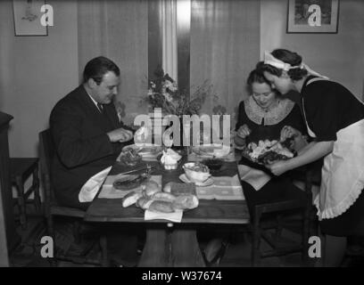 Leben in den 1940er Jahren. Eine junge Frau, die als Dienstmädchen ist, das Abendessen serviert, ein Mann und eine Frau am Tisch sitzen. Sie ist ordentlich in einem weißen Schürze gekleidet. Schweden 1940. Kristoffersson ref 55-7 Stockfoto