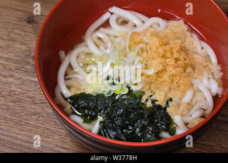 Udon in Brühe mit Wakame Algen, Tenkasu, Frühlingszwiebeln, Fisch Brühe in einen roten Schüssel Stockfoto