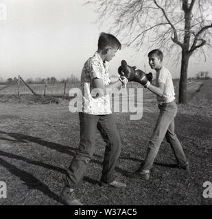 1960er Jahre, historisch, zwei ältere Teenager-Jungen, Brüder, vielleicht 16-18 Jahre alt, in Jeans und kurzärmeligen Hemden und mit Boxhandschuhen, die sich draußen auf einem Feld auf einer Farm, Mid-West, USA, gegenüberstehen. Stockfoto