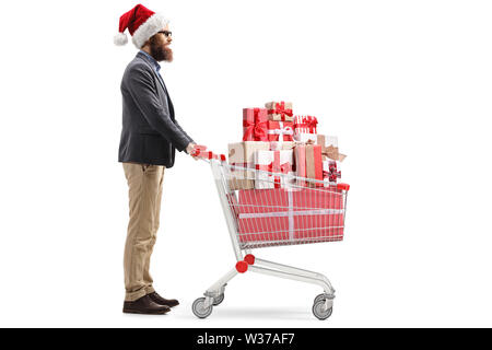 Volle Länge Profil Schuss ein bärtiger Mann mit einem Weihnachtsmann Mütze und stehen mit einem Einkaufswagen voller Weihnachtsgeschenke isoliert auf weißem Stockfoto