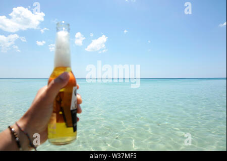 Männliche Hand eine Flasche Bier vor einem sonnigen Himmel und kristallklarem Meer. Ferienhäuser Konzept. Stockfoto