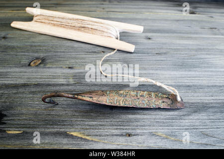 Alte Griff für vertikale blinken Raubfische. Kupfer geschmiedet Spinner und Haspel mit Seil auf Holz- rustikalen Hintergrund. Stockfoto