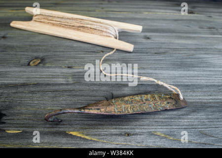 Alte Griff für vertikale blinken Raubfische. Kupfer geschmiedet Spinner und Haspel mit Seil auf Holz- rustikalen Hintergrund. Stockfoto