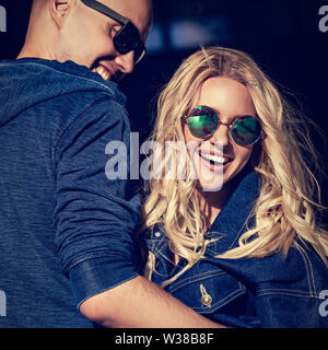 Stilvoll lachen Paar kuscheln in der modernen Blue Jeans Jacken und trendige Sonnenbrille auf Sommer Stadt Hintergrund. Schönen weiblichen blonden Model. Closeup t Stockfoto