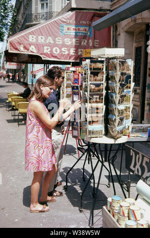 1960, historische, Sommer und zwei Touristen, die auf der Suche nach Postkarten auf einen Stand außerhalb eines Café, Paris, Frankreich. Stockfoto