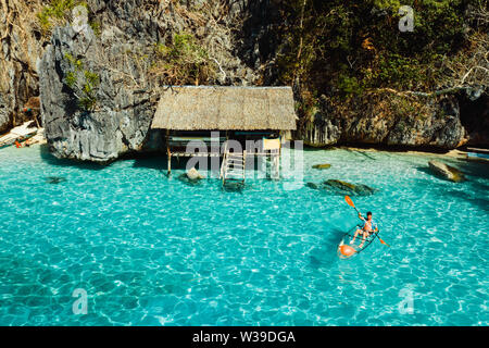 Touristische paddeln auf einem transparenten Kajak an einem tropischen Strand mit blauem Wasser und Palmen - Coron, Philippinen Stockfoto