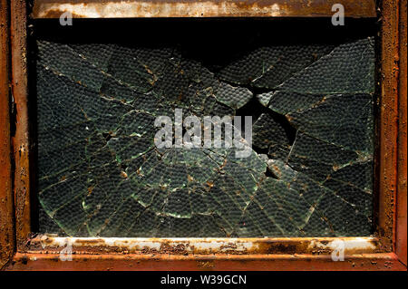 Nahaufnahme eines zerbrochenen, strukturierten Glasfensters, das in einem Rahmen aus rostigem Metall angeordnet ist und radiale Risse und Zerfälle zeigt Stockfoto