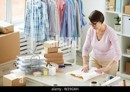Junge brünette Frau Verpackung gefaltet Casualwear in Papier, während im Büro der Online Shop funktioniert Stockfoto