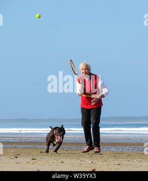 Spaß am Strand, als eine ältere Frau ein Tennisball für Ihren Hund Hits auf dem Sand zu jagen. Stockfoto