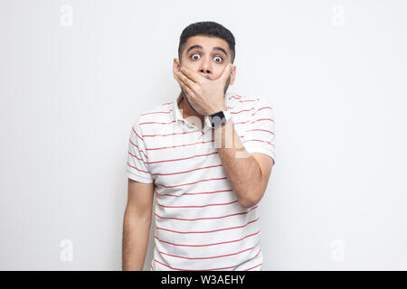 Ich ruhe. Portrait von schockiert, bärtiger junger Mann in gestreiften T-Shirt stand seinen Mund schließen mit Händen und Kamera mit großen Augen. Stockfoto