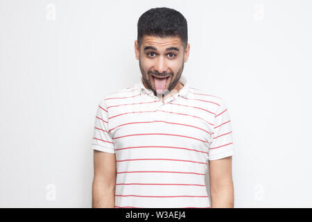 Portrait von Lustig verrückt aussehenden Bärtigen jungen Mann in gestreiften T-Shirt stehen, Zunge raus, und wenn man die Kamera mit lustigen Gesicht. indoor Studio shot, Stockfoto
