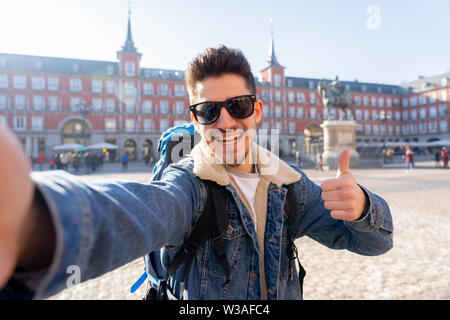 Attraktive junge touristische student Mann mit Rucksack, Spaß haben, glücklich und aufgeregt, wobei ein lächelndes selfie mit Smart Phone an der Plaza Mayor, Madrid, Spanien Stockfoto