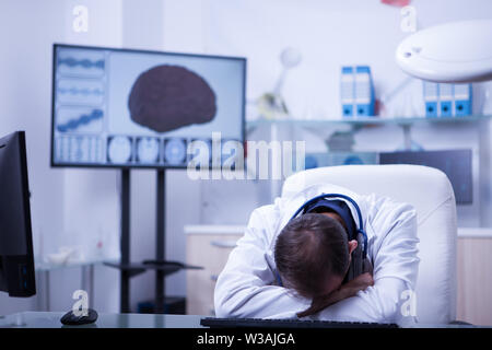 Junge kaukasier Arzt an diesem Schreibtisch sitzen im Krankenhaus mit seinen HEAND auf dem Schreibtisch schlafen. Overwokerd Arzt. Stockfoto