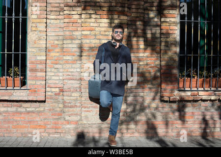 Lifestyle outdoor geschossen von Stattlichen fröhliche junge Mann Tasche über der Schulter und wartet, Freund, gegen die Mauer im Hintergrund stehend Stockfoto