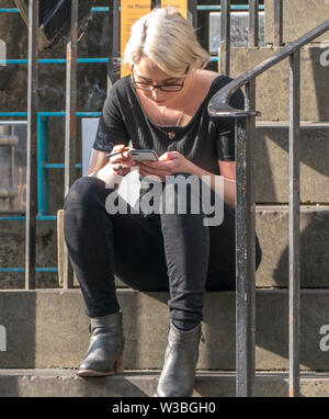 Blonde junge Frau eine Pause, sitzen auf den Steinstufen draußen in der warmen Sonne, rauchen eine Zigarette, während Ihr Handy suchen. Stockfoto
