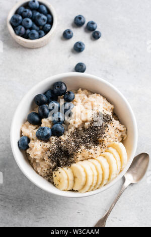 Lecker brei Haferflocken mit Bananen Blaubeeren und Chia Samen in Weiß Schüssel, Table Top anzeigen. Gesundes Frühstück zu essen. Grauer Beton Hintergrund Stockfoto