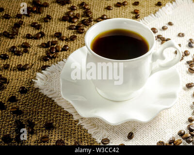 Heißer Schwarzer Kaffee in einem weißen elegante Schale auf über Hintergrund in beige Tabelle Serviette mit Fransen und Kaffeebohnen. Morgen heißen Getränk. Kaffee Stimmung und l Stockfoto