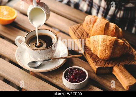 Gießen Creme in Tasse schwarzen Kaffee. Leckeres Frühstück mit Croissants, Marmelade und Kaffee Stockfoto