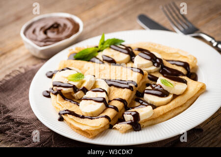 Crêpes mit Schokoladensauce und Bananen auf weiße Platte, hölzernen Tisch Hintergrund Stockfoto