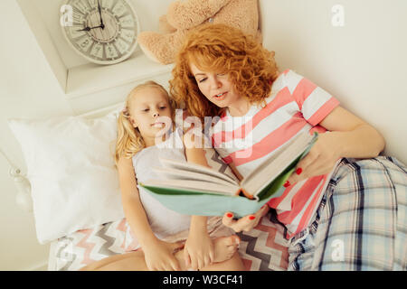 Gute-nacht-Märchen. Blick von oben auf eine angenehme schöne Frau mit einem Buch beim zusammen mit ihrer Tochter liegen Stockfoto