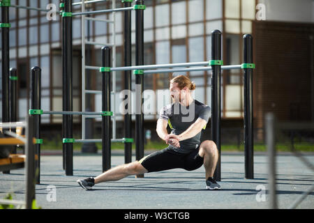 Ernsthafte schlanker junger Mann in enger Kleidung üben Adduktoren stretch Übung auf Outdoor Training Boden in der Stadt Stockfoto