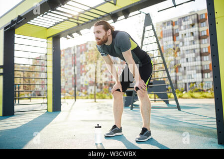 Müde junge bärtiger Mann mit Bart stützte sich auf die Knie, während sie Atem nach intensiver Übung auf Sportplatz Stockfoto