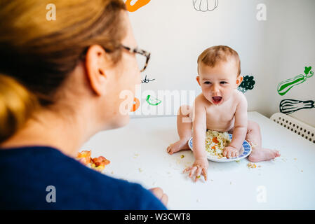 Baby glücklich lächelnd auf seine Mutter sah ihn beim Essen mit den Händen in die Küche. Stockfoto