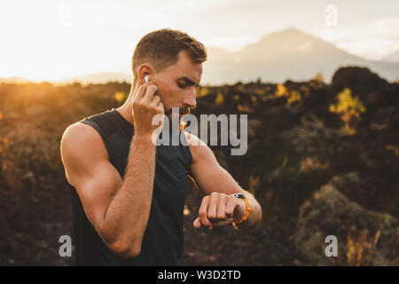 Männliche Läufer Synchronisierung Drahtloser Ohrhörer mit smart Watch. Vorbereitung für Trail Running im Freien bei Sonnenaufgang. Stockfoto