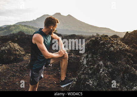 Junge hipster Runner mit Bart dehnen und Aufwärmen für Trail Running im Freien. Musik hören in der Luft Pods. Blick auf die Berge im Hintergrund. Stockfoto