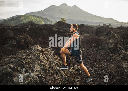 Junge hipster Runner mit Bart dehnen und Aufwärmen für Trail Running im Freien. Musik hören in der Luft Pods. Blick auf die Berge im Hintergrund. Stockfoto