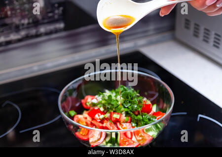 Vorbereitung von Salat von frischem Gemüse, Gurken, Tomaten und grün mit einem weißen Plastiklöffel aus dem gießt aus einem Dressing aus Olivenöl, Balsam Stockfoto