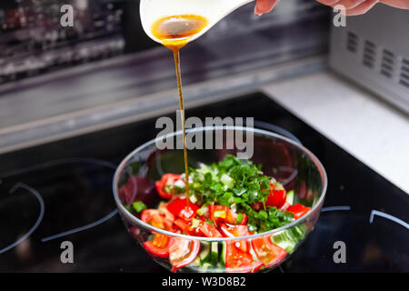 Vorbereitung von Salat von frischem Gemüse, Gurken, Tomaten und grün mit einem weißen Plastiklöffel aus dem gießt aus einem Dressing aus Olivenöl, Balsam Stockfoto