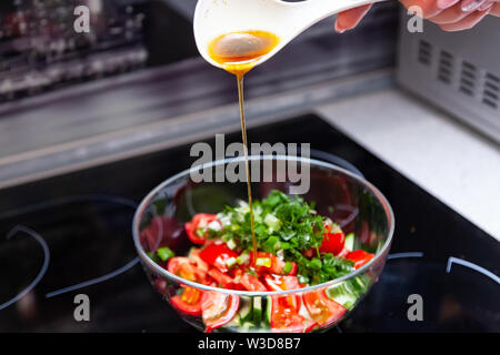 Vorbereitung von Salat von frischem Gemüse, Gurken, Tomaten und grün mit einem weißen Plastiklöffel aus dem gießt aus einem Dressing aus Olivenöl, Balsam Stockfoto