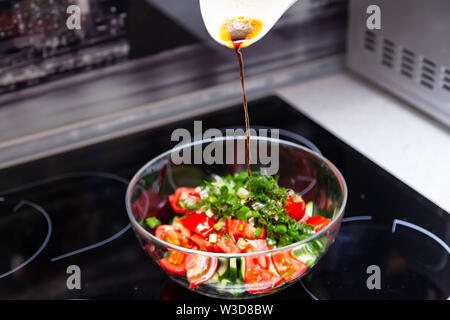 Vorbereitung von Salat von frischem Gemüse, Gurken, Tomaten und grün mit einem weißen Plastiklöffel aus dem gießt aus einem Dressing aus Olivenöl, Balsam Stockfoto