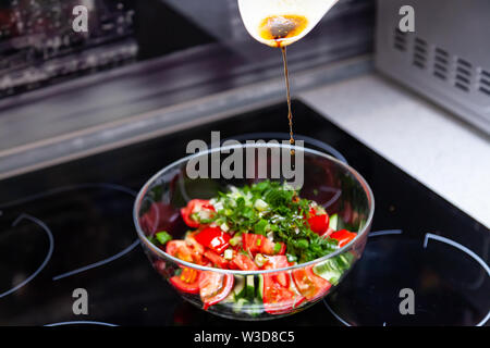 Vorbereitung von Salat von frischem Gemüse, Gurken, Tomaten und grün mit einem weißen Plastiklöffel aus dem gießt aus einem Dressing aus Olivenöl, Balsam Stockfoto