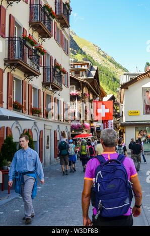 Zermatt, Schweiz - 10. Juli 2019: Touristen zu Fuß auf der Straße von Alpine Village Zermatt in der Sommersaison. Wanderung, Wanderer. Schweizer Alpen. Kleinen Bergstadt. Schweizer Fahne im Hintergrund. Stockfoto