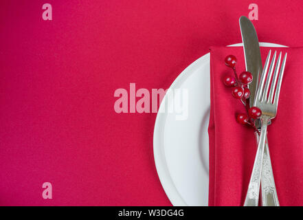 Silber Messer und Gabel, rot Bettwäsche Serviette und roten Stechpalme Beeren liegen auf dem weißen Porzellan Teller, die auf einem Tisch mit einem roten Tabellec befindet. Stockfoto