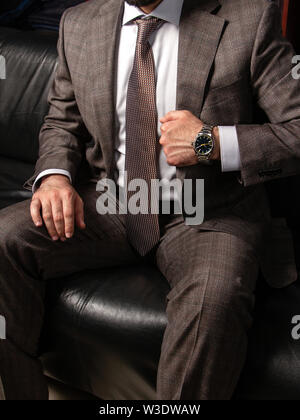 Ein junger Mann in einem klassischen Anzug und teure Armbanduhr sitzt in einer angespannten Haltung auf ein schwarzes Ledersofa. Style gesichert Herren. Demonstration von excelle Stockfoto