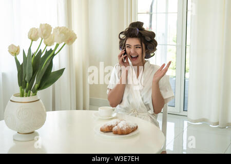 Schöne junge lächelnde brünette Frau in Lockenstäbe sprechen auf dem Smartphone beim trinken Kaffee und essen Croissants zum Frühstück im hellen Stockfoto
