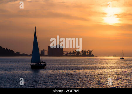 Segelboot bei Sonnenuntergang auf dem Fluss Tejo vor Porto Brandao Hafen und der industriellen Zone, Lissabon, Portugal Stockfoto