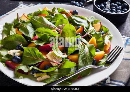 Nahaufnahme der Salat mit frischen Mangold Blätter, Pfirsich, Blaubeeren, Stücke von blauen Schimmel Käse serviert auf einem weißen Teller auf einen konkreten Tisch mit Zutaten Stockfoto