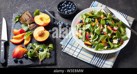 Salat mit frischen Mangold Blätter, Pfirsich, Blaubeeren, Stücke von Gorgonzola Käse auf einem weißen Teller auf einen konkreten Tisch mit Zutaten serviert, Schmierfilm Stockfoto
