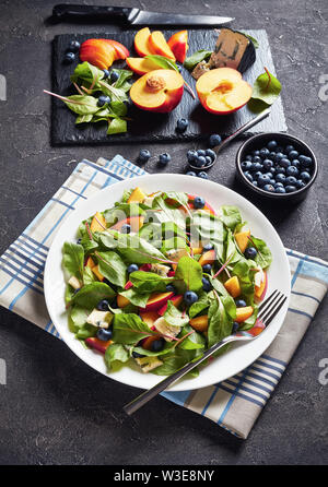 Salat mit frischen Mangold Blätter, Pfirsich, Blaubeeren, Stücke von Gorgonzola Käse auf einem weißen Teller auf einen konkreten Tisch mit Zutaten serviert, vertikal Stockfoto